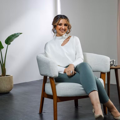 Taiba Al-humaidhi sitting on a bench, wearing a white shirt and black pants, smiling to the camera